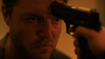 Legends TV show screenshot 1: Tom Burke presses his forehead against the end of a handgun. He wears a collared shirt.