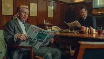 Legends TV show screenshot 2: Steve Coogan and Douglas Hodge sit at a desk reading newspapers. Coogan wears a gray suit, Hodge wears a black suit.