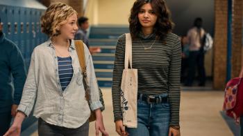 Two students walk down a hallway lined with blue lockers. The blonde student speaks to the brunette student while gesturing with her hands.