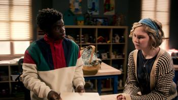 Two students (one a black boy in a colorful pullover, one a blonde girl in a tan striped cardigan) stand at a table inside a classroom, the girl appears to be listening to something the boy is saying. Her brow is slightly furrowed.