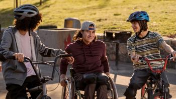 Three boys are pictured outside in a paved area in front of a grassy hill. Two are wearing bike helmets and sitting on bikes, while the middle boy sits in a wheelchair wearing a backwards baseball cap. They are smiling at one another.