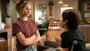 A blonde girl in a red t-shirt crosses her arms and looks down at a shorter boy with brown hair who is on crutches. She has a skeptical look on her face. They are standing near the kitchen inside a suburban home.