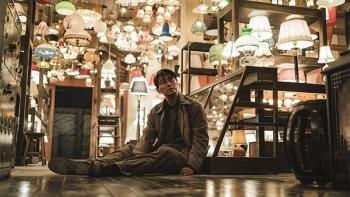 Light Shop TV show screenshot 2: A man sits on the floor of a brightly-lit light shop, various lamps and lighting fixtures hanging around him