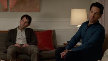 Living with Yourself Series: The two Mileses sit together in a living room, craning their necks to see something off-screen. 