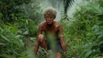 Lord of the Flies TV show screenshot 1: Jack crouches in between two bushes, wearing only shorts; he stares into the distance with wide eyes