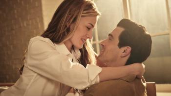 Love Story TV show screenshot 2: Sarah Pidgeon and Paul Anthony Kelly lay on a chair. Pidgeon wears a white top, while Kelly wears a brown shirt.