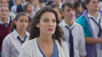 Love 101 TV screenshot 2: Burcu wears a blue top and white cardigan, a group of students behind her in softer focus