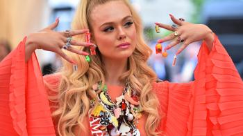 Elle Fanning wears a coral kaftan blouse with lots of jewelry. She holds up her hands, showing pink painted nails with charms.