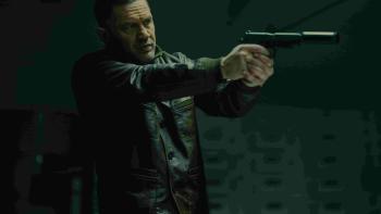 Tom Hardy stands in a dark room, aiming a gun with a silencer. He wears a dark leather jacket.