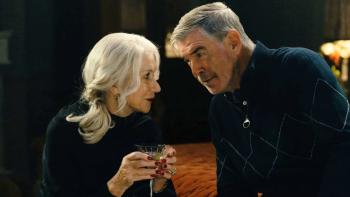 (From L-R) Helen Mirren and Pierce Brosnan sit in a room together. Mirren holds a glass of wine. She wears a black sweater.