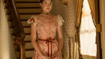 The Lizzie Borden Story screenshot 2: Ella Beatty stands at the bottom of a staircase., wearing a light pink floral dress.