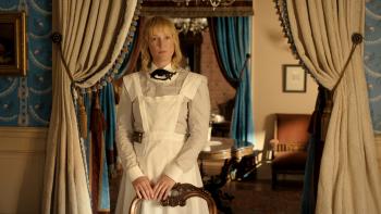 The Lizzie Borden Story screenshot 1: Vicky Krieps wears a light grey dress, with a bow at the collar and apron. She rests her hands on a chair.