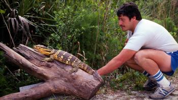 Monsters of God: Man crouching beside a large lizard on a log in a tropical setting.