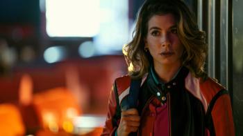 Giulia Michelini wears a red, white, and black leather racing jacket, and a red buttoned blouse. She holds a black bag strap.