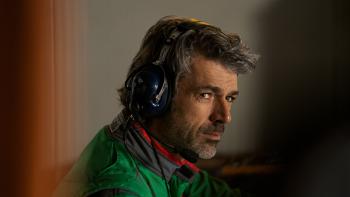Luca Argentero wears a green, black, and red trimmed pit crew racing jacket. He wears large dark blue over ear headphones.