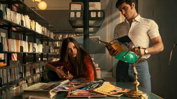 Mrs. Playmen TV show screenshot 1: Elsa and Steve are shown at a desk inside a library, thumbing through news magazines