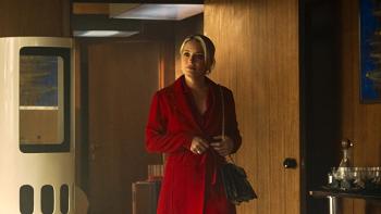 Mrs. Playmen TV show screenshot 2: Adelina wears a red coat and stands in the doorway of a wood-paneled office, a bar cart to her left