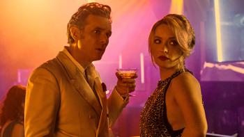 Mrs. Playmen TV show screenshot 3: Saro and Adelina stand together in a colorfully-lit club, Saro holds a drink; Adelina glances over her shoulder