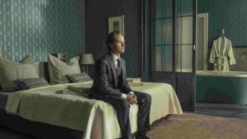 Murder Mindfully TV show screenshot 1: Tom Schilling sits on a bed in a blue hotel room. He wears a dark blue suit and black shoes.