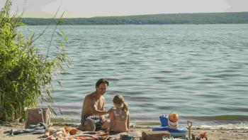 Murder Mindfully TV show screenshot 2: Schilling and Pamuk Pilavci sit on the sand in front of a lake. They're surrounded by toys, towels, and bags.