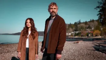 A man and woman in coats stand on a rocky beach looking off in the distance with town in the distance behind them.