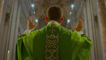 Mysteries of the Faith: The back of a priest in a bright green robe is shown as he stands in the front of a church with his hands raised during a sermon.