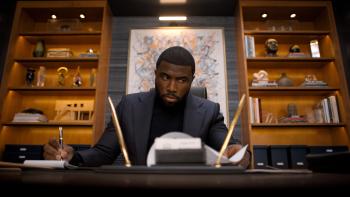    ss-2: Y’lan Noel sits at a desk, writing. He wears a black suit jacket and black turtleneck.