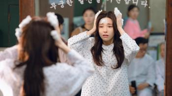 Ha-young holds bandaged fingers against her head as she looks into a mirror. She wears a white hospital gown.