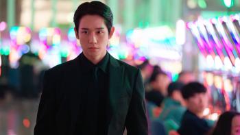 Jung Hae-in walks through a room with bright lights. He wears a suit, all-black.