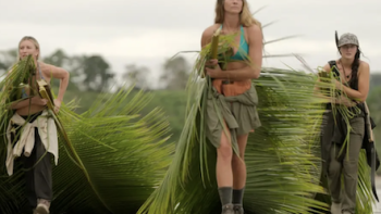 Outlast: The Jungle TV show screenshot 3: Three women carry large palm fronds on a cloudy day.