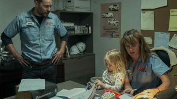 Painkiller TV Show Screenshot 2: Taylor Kitsch stands over Carolina Bartczak sitting at a desk. Carolina has a child in her arms, and they both wear b