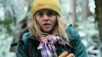 Penelope stands in the woods looking cold, her nose slightly pink. She wears a green coat and yellow hat with blood on it