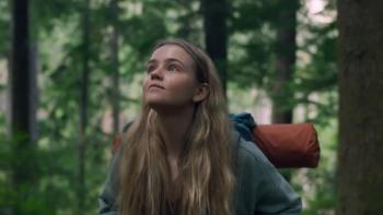Penelope stands in the forest, gazing up with an appreciative look on her face. She wears an orange pack on her back