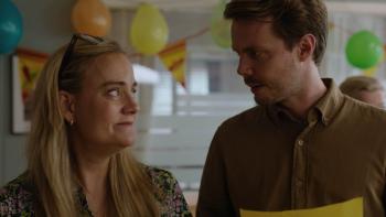 Pernille TV show screenshot 1: A middle aged woman, and slightly younger man look at each other with closed lip smiles in a room full of balloons
