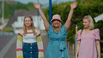 Pernille TV show screenshot 2: Pernille stands between her teen daughters in a teal dress with arms in the air joyfully waving while the teens scowl