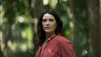 Morgana O'Reilly stands in a forest. She wears a red button-down shirt with thin white stripes.
