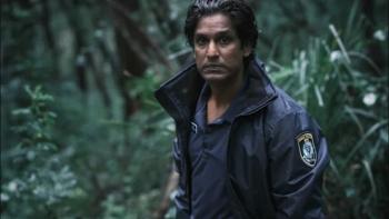 Rudi Dharmalingam stands in a forest. He wears a black jacket with a police patch on the arm, and a dark shirt with a name tag.