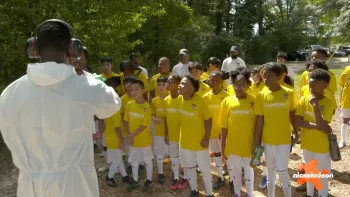 Play It Forward TV show screenshot 1: A diverse group of kids wearing a yellow Gameday shirt listen attentively to a man in white coveralls.