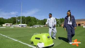 Play It Forward TV show screenshot 3: Kel Mitchel and Renee Montgomery watch a field liner robot draw white lines on a soccer field.