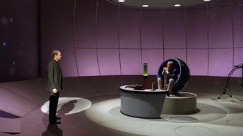 The Prisoner TV show screenshot 2: A large, rounded Space Age-style room with purple ombre walls houses a curved desk and egg-shaped chair. A man stands at the left of the image, speaking to another man who sits in the chair.