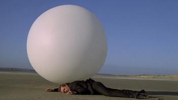 The Prisoner TV show screenshot 3: A fully clothed man lays unconscious, face-down on a desolate beach with a huge white orb resting upon his back.