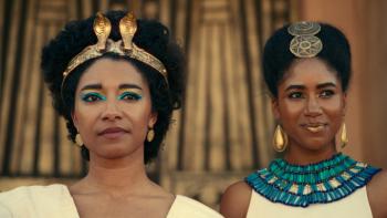 Queen Cleopatra TV show: Cleopatra and Arsinoe are shown onstage during a royal celebration with faces in closeup. Both wear elaborate hairstyles; Arsinoe ha a beaded turquoise collar and Cleopatra has a gold crown with snakes. 