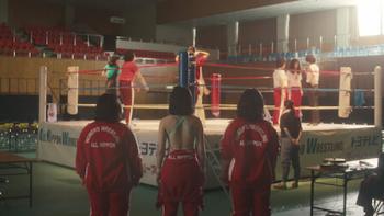 The Queen of Villains screenshot 2: Three aspiring female wrestlers, pictured from behind, stand in a gym watching others work inside the ring
