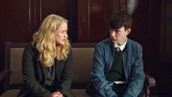 Blonde middle aged woman seated on a bench looking earnestly at a teenage brunette boy to her left.