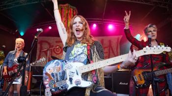 Riot Women TV show screenshot 1: Kitty is center-stage holding a guitar and raising her arm in triumph; Jess and Holly flank her, holding instruments