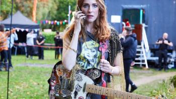 Riot Women TV show screenshot 3: Kitty wears her guitar, holds a phone to her ear and looks horrified; behind her, workers set up for a music festival