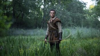 Jack Patten stands in a foggy field of grass. He wears a brown tunic and leather gloves. He carries a bow.