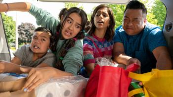 Run the Burbs TV show screenshot 2: All four of the Phams are looking into a car's trunk stuffed with groceries. Leo grimaces with effort; Khia and Andrew are both attempting to pick up heavy bags. 