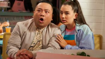Run the Burbs TV show screenshot 3: Khia has her arm around Andrew as the two sit together at a dining room table. Andrew's mouth is open, seemingly in shock. 