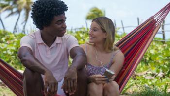 Saint X TV show: Alison and Edwin sit together on a hammock. She's in a bathing suit, he's in his resort work uniform. They look at each other flirtatiously. 